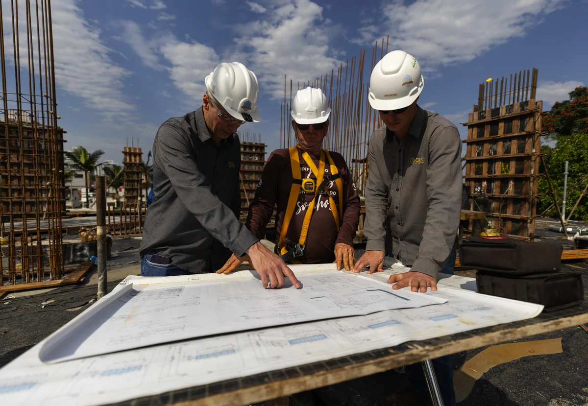Técnico de segurança e engenheiros da CILL analisam o projeto no canteiro de obras, todos com capacete de proteção